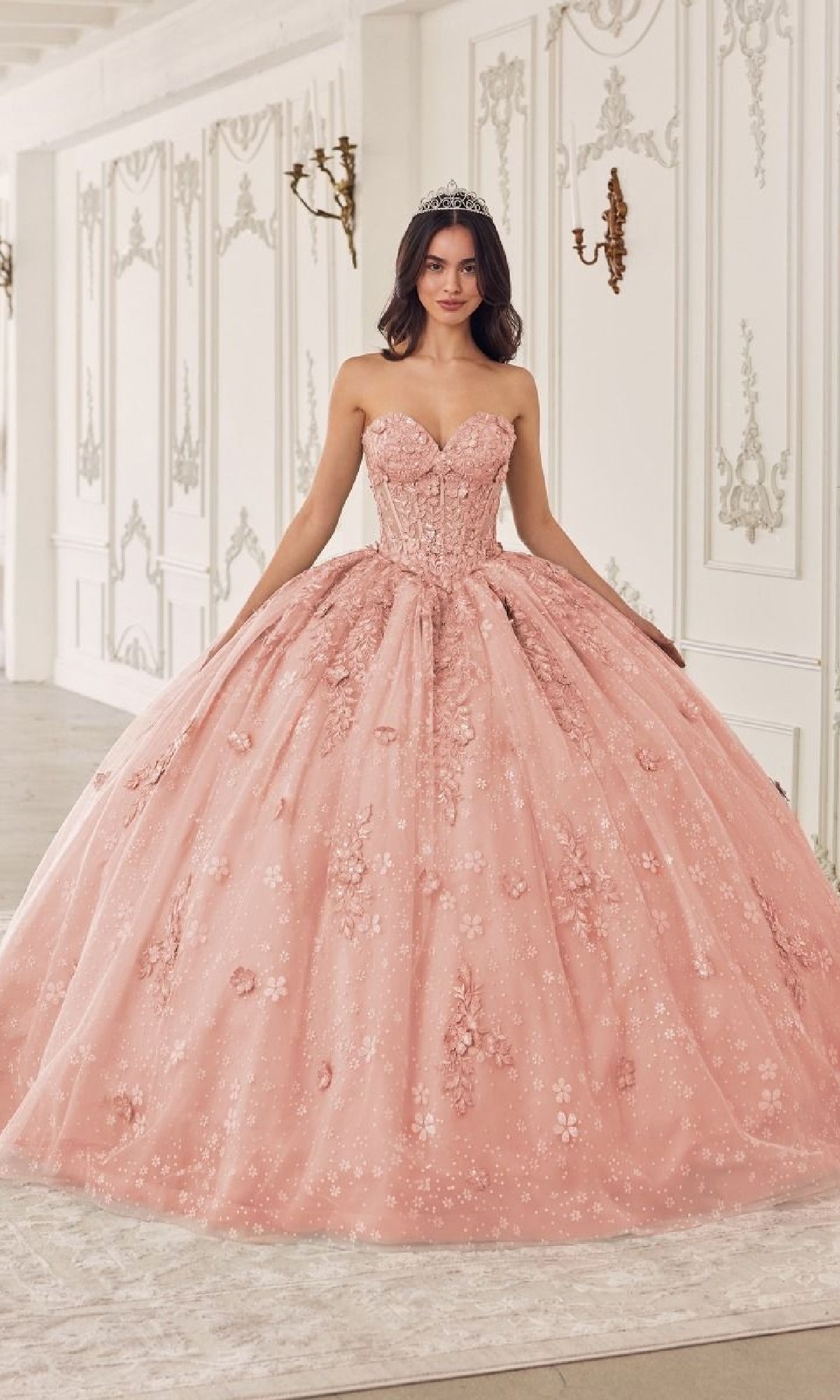 A woman stands in an ornate room with white and gold decor, wearing the Quinceanera Dress 15722 by Ladivine—a pink ball gown with floral details and removable puff sleeves—her tiara adding a regal touch.
